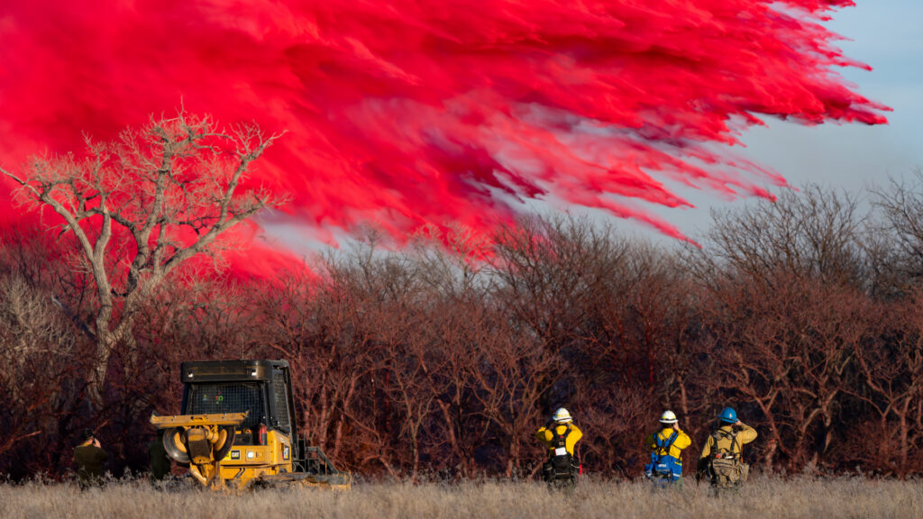 Helicopter drops red fire retardant over a wildfire area while firefighters watch from the ground.