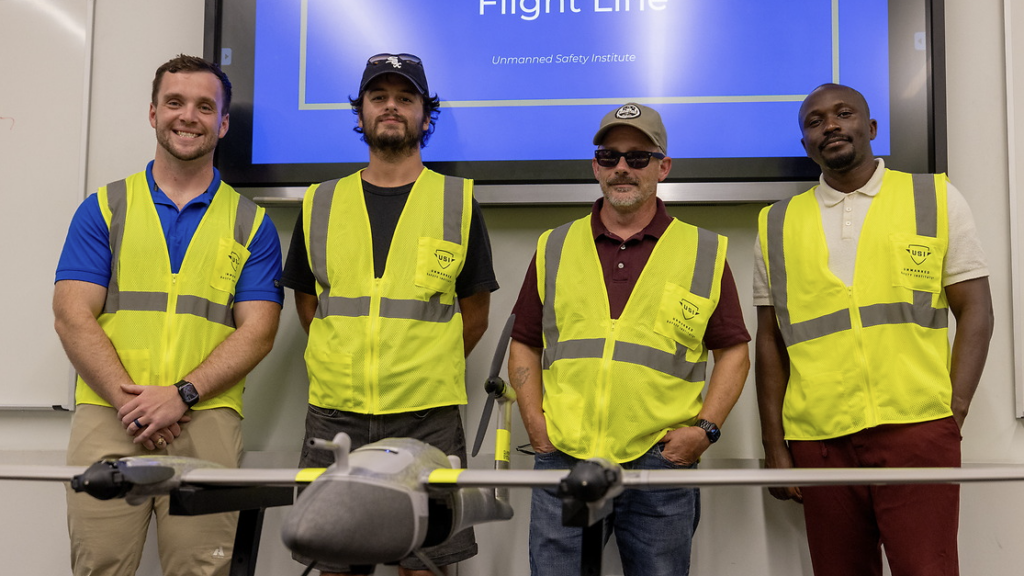 Four people wearing high-visibility safety vests stand behind a fixed-wing drone displayed on a table in a classroom or training room. A presentation screen is visible in the background.