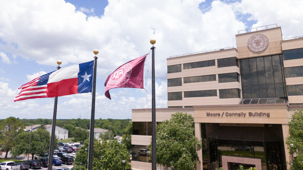 Three flags, including the United States flag, Texas flag and Texas A&M flag, fly in front of The Texas A&M University System's Moore/Connally Building in College Station, Texas, under a partly cloudy sky.