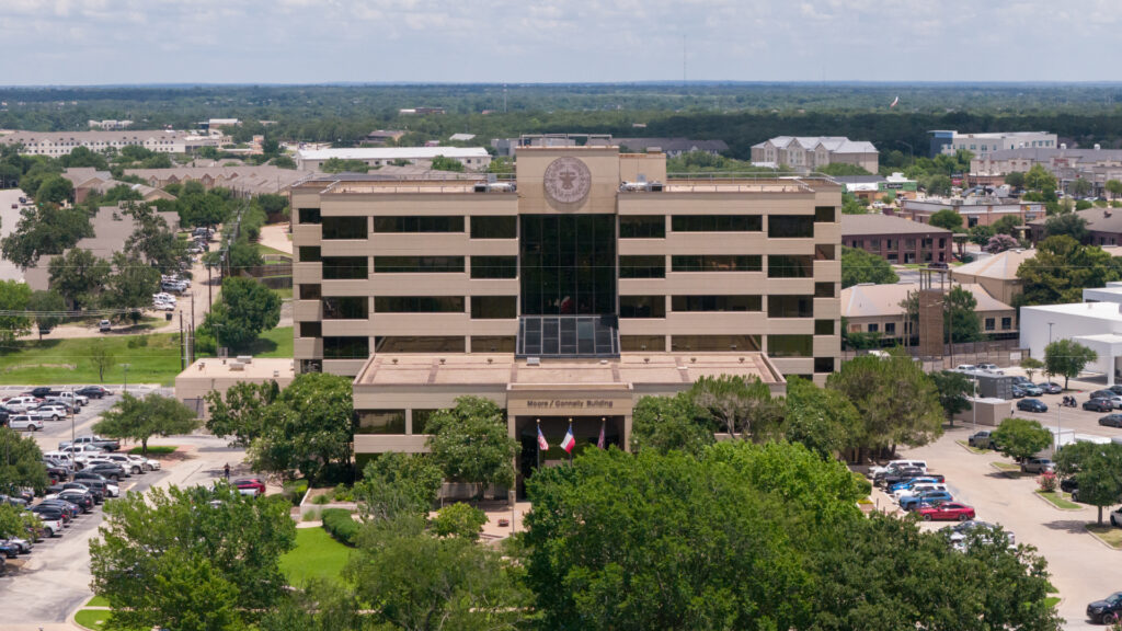Aerial view of the Moore/Connally Building, headquarters of The Texas A&M University System, surrounded by trees, parking lots and commercial buildings in College Station, Texas.