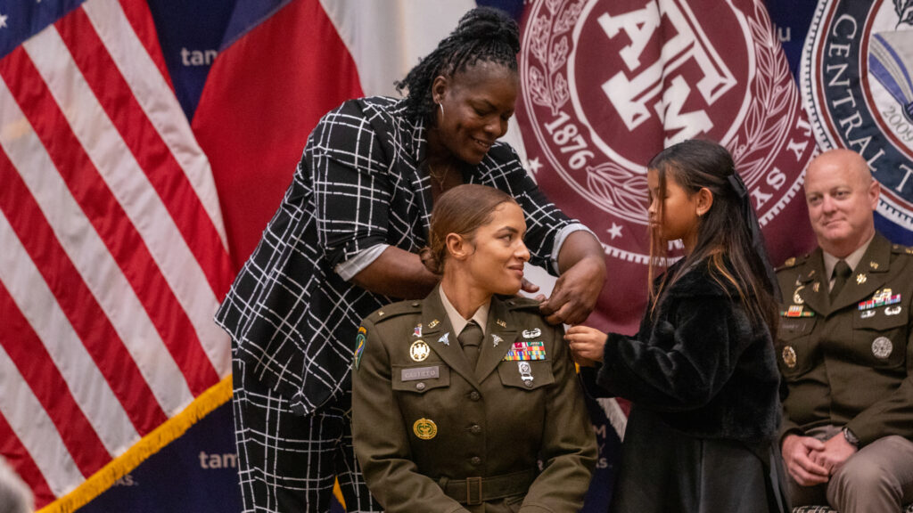 A military promotion ceremony takes place in front of Texas A&M University System banners and U.S. and Texas flags. A uniformed service member sits while two family members assist with attaching new rank insignia. Another uniformed service member is seated in the background.