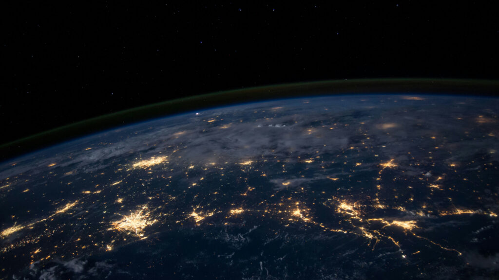 View of Earth from orbit at night, showing illuminated city lights, cloud cover and the planet's curved horizon against the darkness of space.
